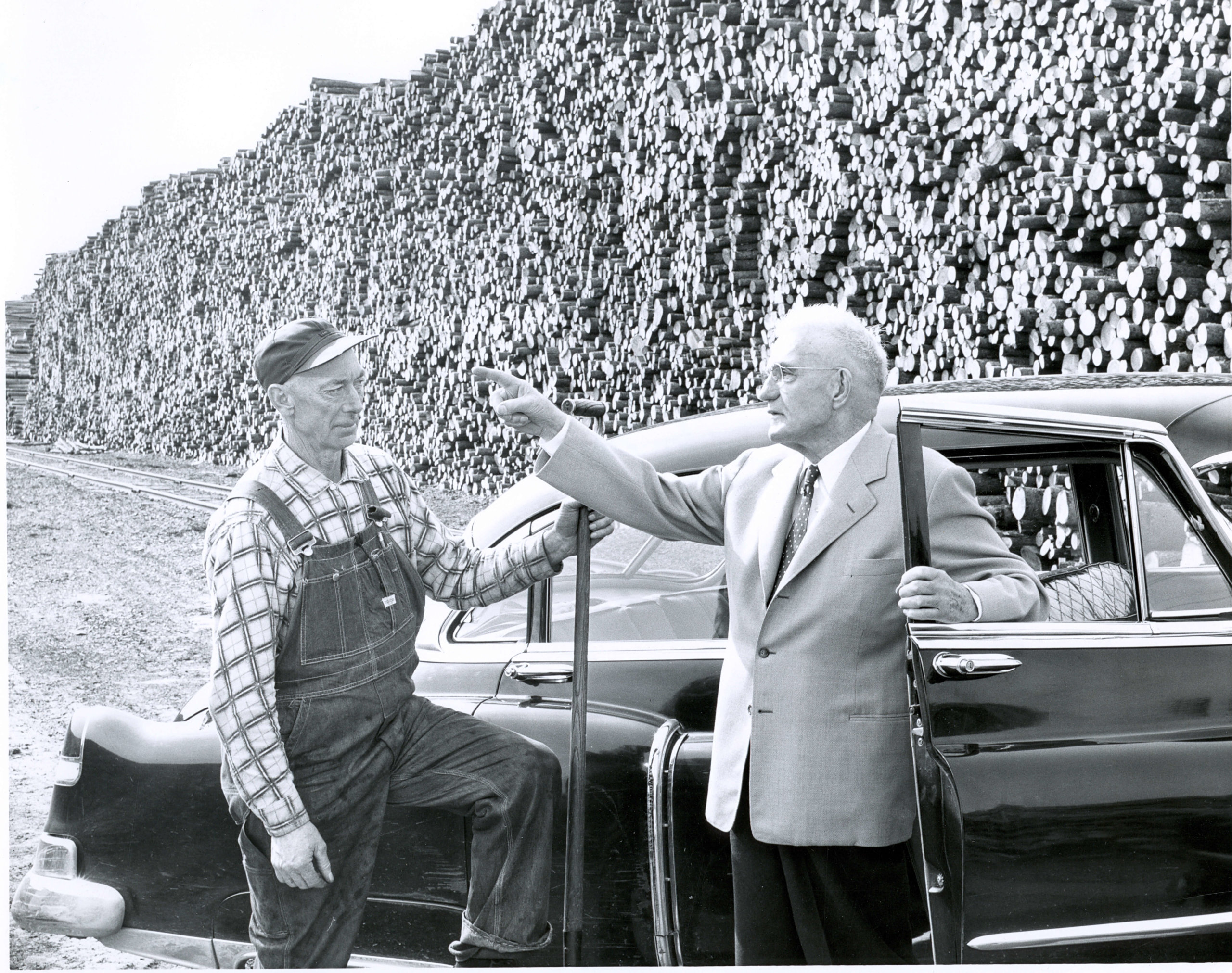 CK by car talking to man in overalls log pile in bkgd 1950.364.10001