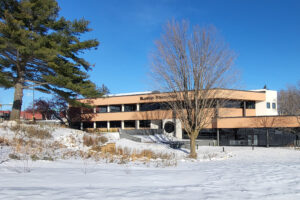 Photo of the Blandin Foundation office as seen from the river, with snow covering the ground, a bright blue sky, and a mature white pine to the left.