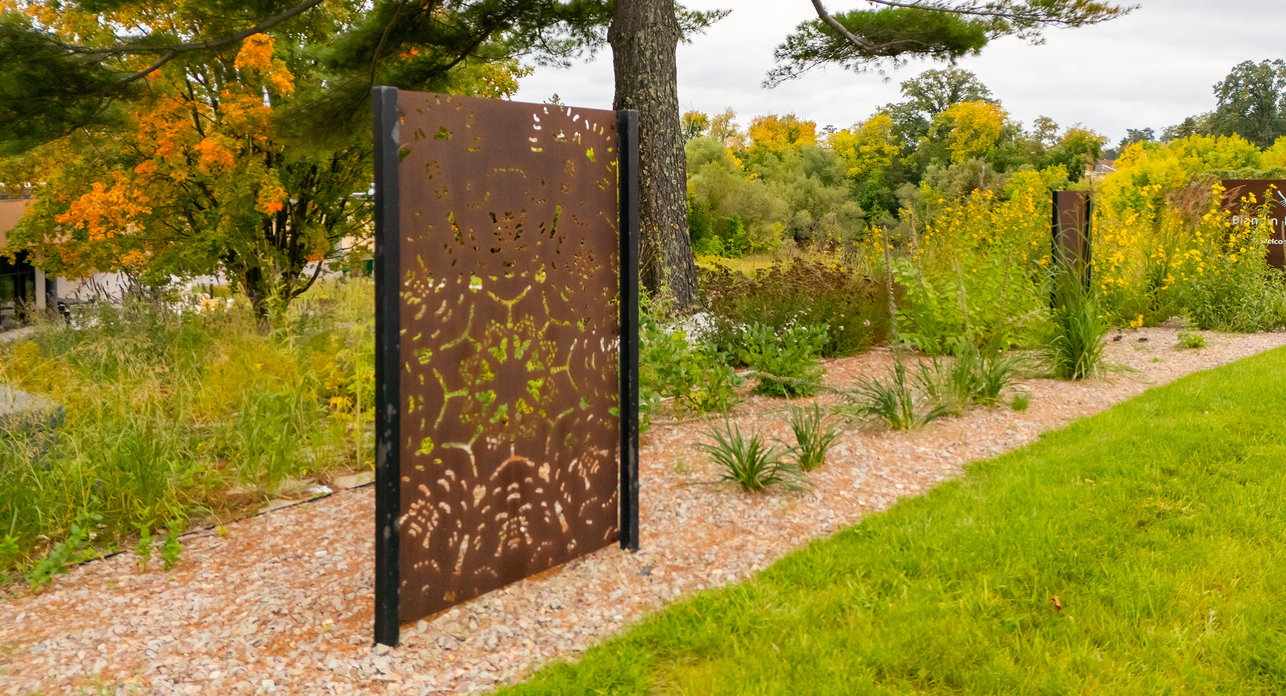 A metal panel with cutouts for a birch bark biting pattern radiating from a center point and symmetrical across four axes, on a stone path with grass, gardens and trees around.
