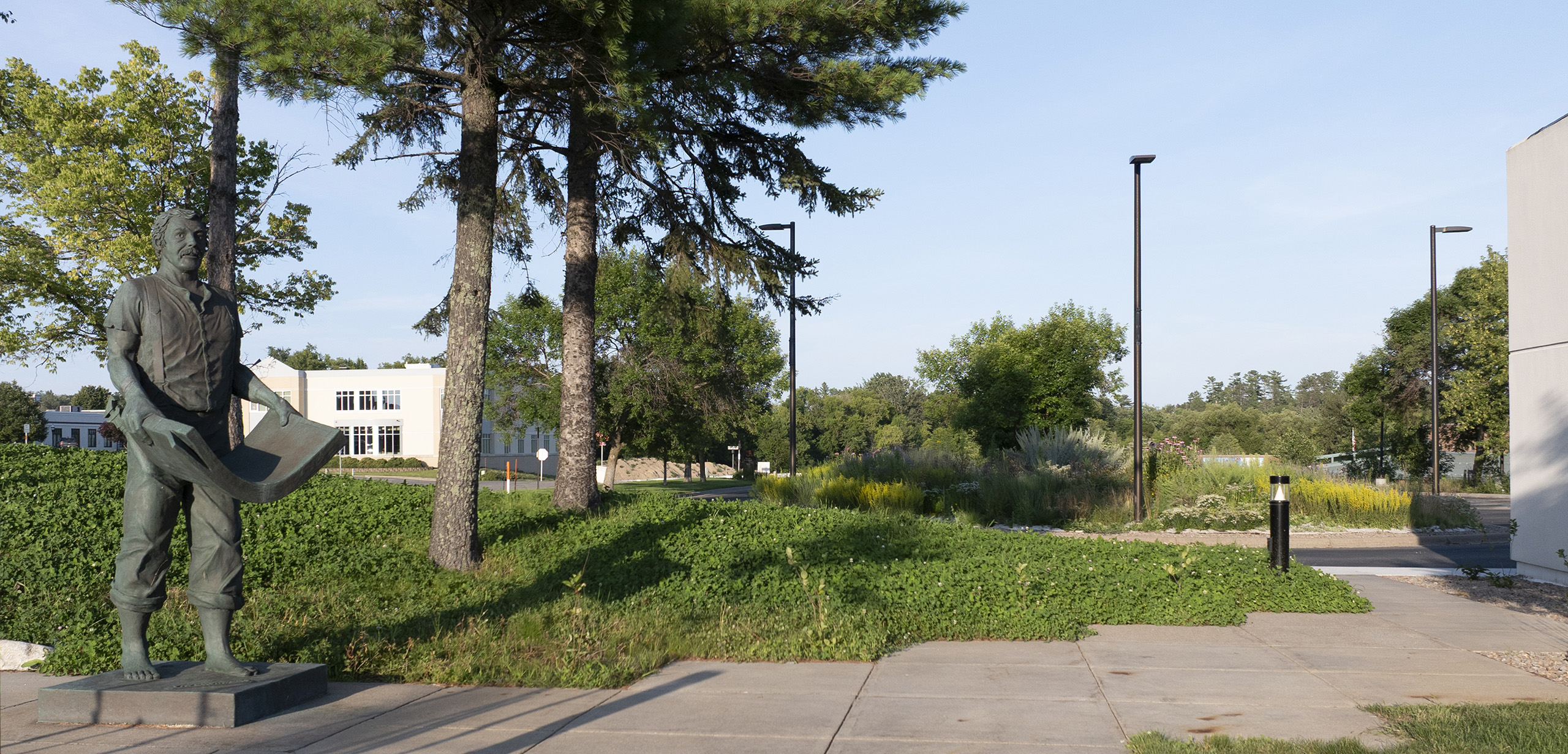 Statue of a working man holding a thick stack of large paper bowing toward the ground, standing outside the Blandin Foundation office with trees and a clear sky in the background.